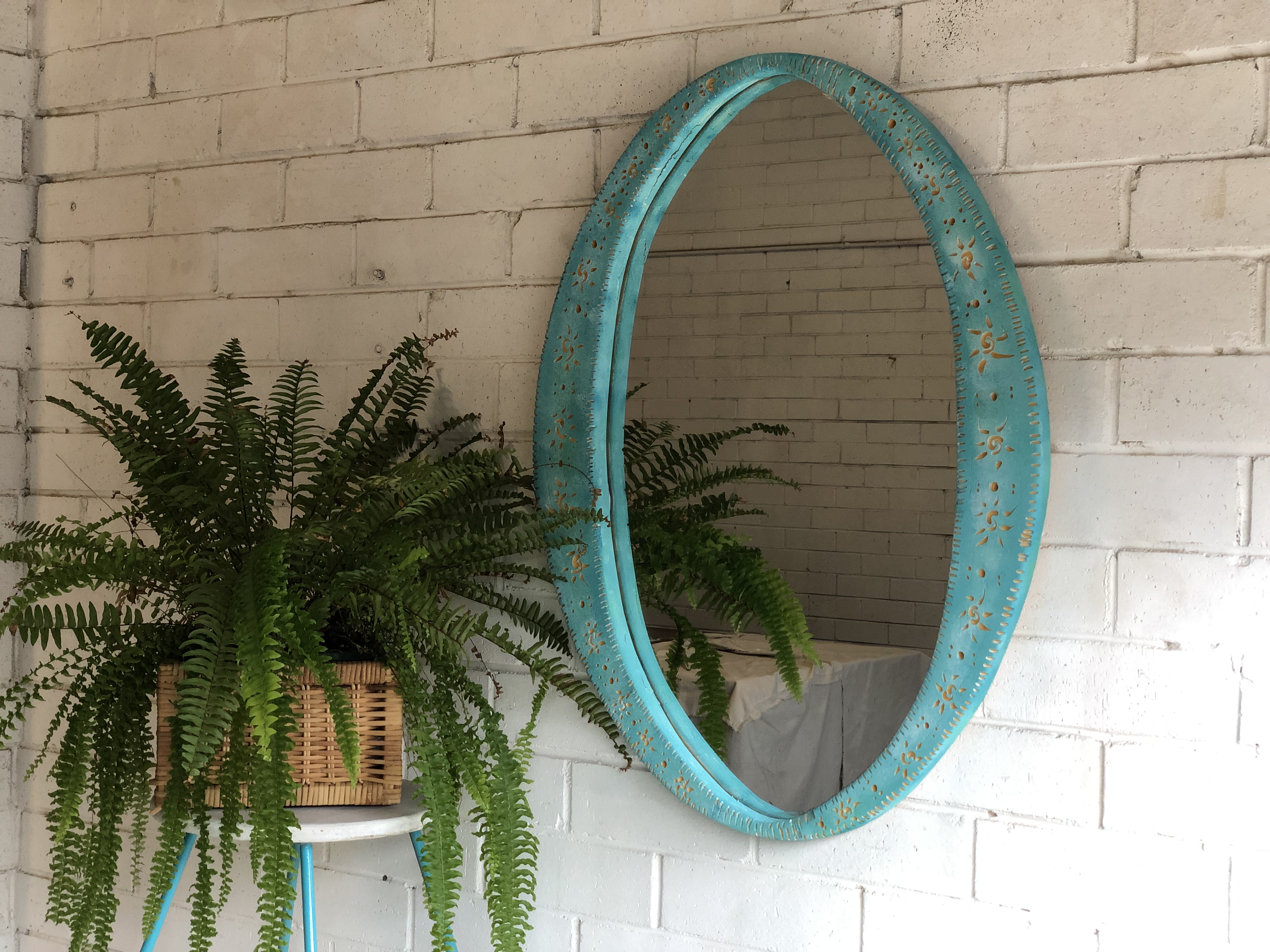 On a white, brick wall hangs a mirror with an ornate blue frame. On the left a fern sits on top of a stool