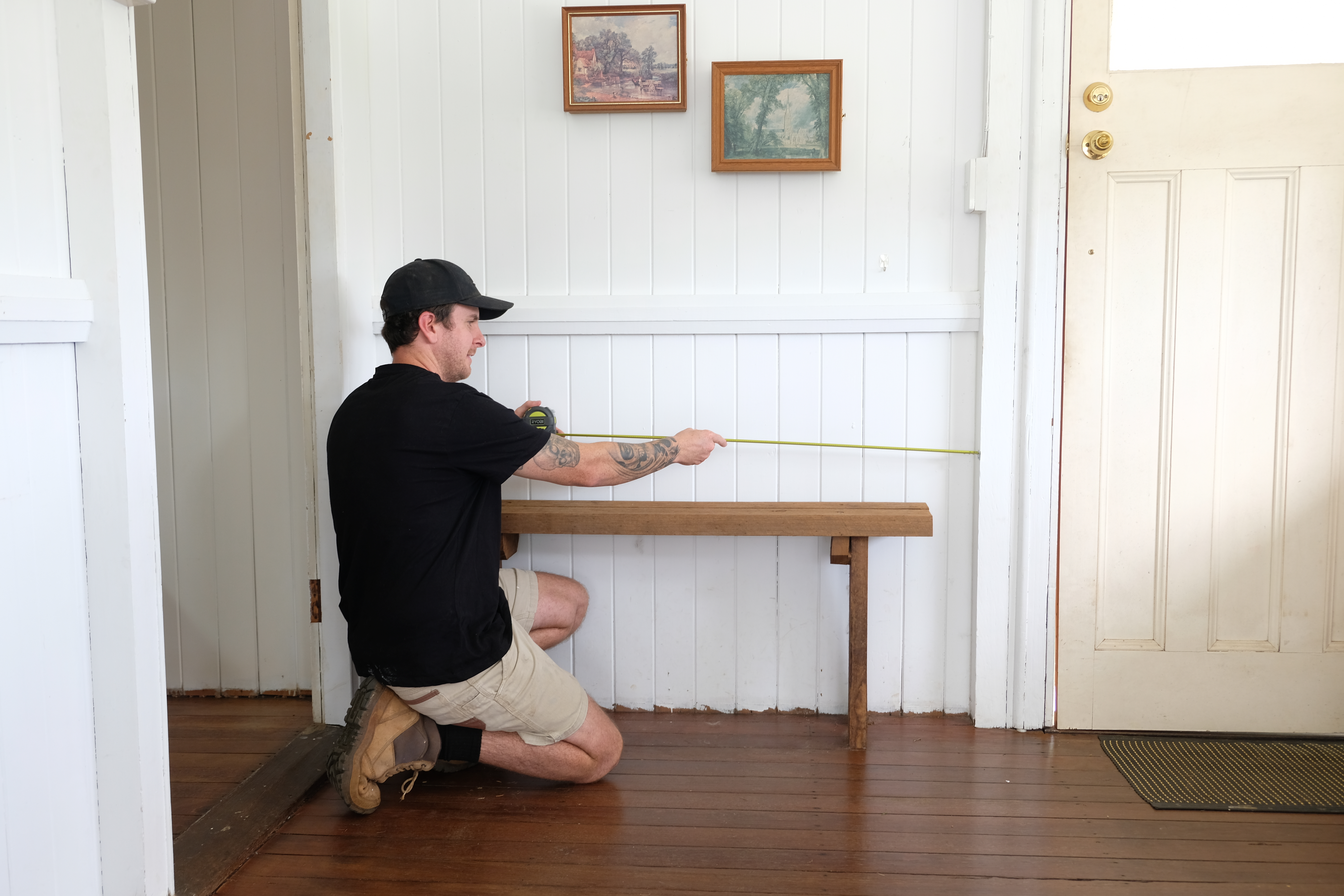 Jake uses a RYOBI measuring tape to plan the size of the DIY shoe cabinet. The space he's chosen contains a bench and has photo frames on display.