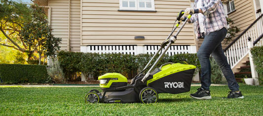 A man using a RYOBI lawn mower to cut the front lawn of a residential property. The lawn is well-maintained and freshly cut. 