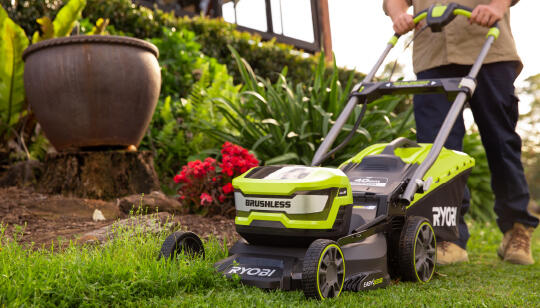A man using a RYOBI lawn mower being using to cut a patch of overcut grass next to a garden bed. 