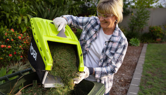 A woman wearing safety glasses and gloves, emptying a catcher full of grass clippings (from a RYOBI lawn mower) into the bin. 