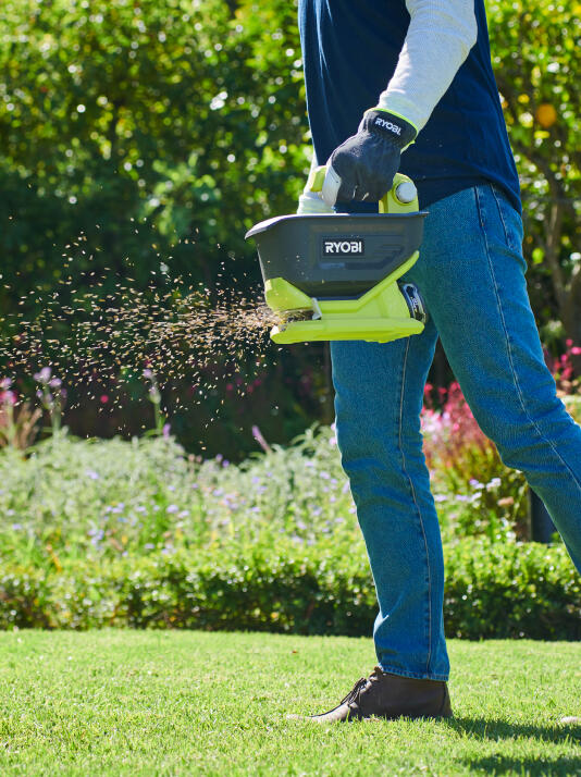 A person wearing gloves, using a RYOBI seed &amp; fertiliser spreader to distribute fertiliser onto a well-maintained lawn. 